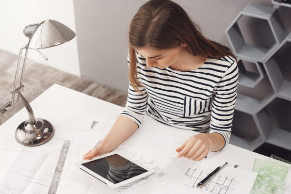 Job, career and business concept. Portrait of young fashionable professional female designer sitting at table, looking in digital tablet monitor, chatting with customer to decide some details